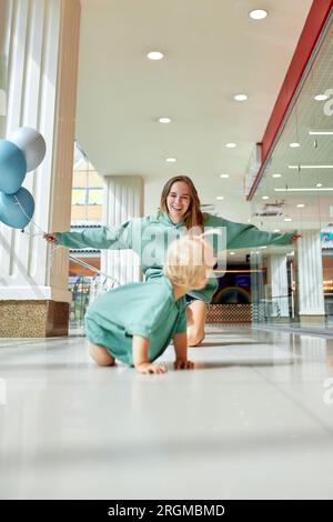 Felice madre sorridente con palloncini colorati e il suo bambino gattonava sul pavimento. La giovane bella mamma e la sua piccola figlia in abiti verdi camminano intorno a me Foto Stock