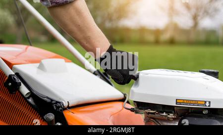 L'uomo irriconoscibile del giardiniere nei guanti di protezione avvia il rasaerba prima di tagliare il prato verde nel suo cortile. Uomo con rasaerba motorizzato ca Foto Stock