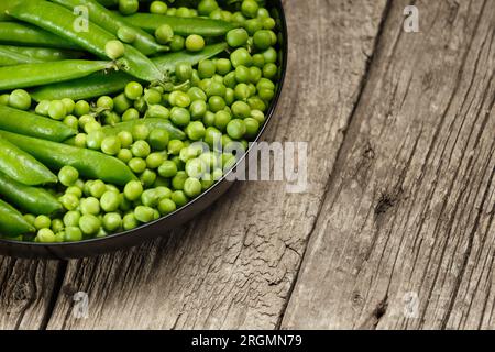 Cialde di piselli verdi e piselli freschi biologici sgusciati in un piatto nero rotondo su uno sfondo di legno invecchiato, spazio copia. Proteine vegetali, prodotti sani. Foto Stock