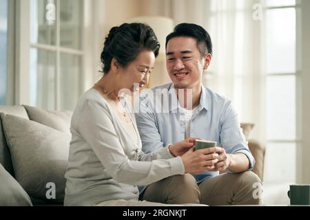 figlio asiatico adulto seduto sul divano a casa chiacchierando con la madre anziana felice e sorridente Foto Stock