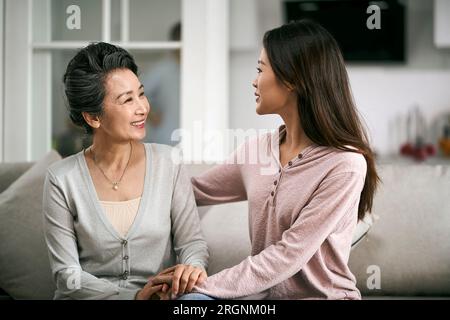 figlia asiatica adulta seduta sul divano a casa chiacchierando con la madre anziana felice e sorridente Foto Stock