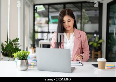 Una bella donna d'affari asiatica si concentra sui suoi compiti, esaminando i rapporti finanziari alla sua scrivania in ufficio. Foto Stock