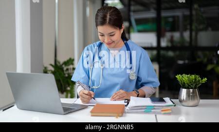 Una bella e professionale medico asiatico in scrub che lavora sui suoi casi medici e legge una ricerca medica sul suo portatile nel suo ufficio a. Foto Stock
