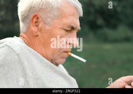 Uomo anziano che accende una sigaretta all'aperto su sfondo verde. Vecchio uomo premuroso che fuma, guardando da parte. Faccia da vicino. Pensione rilassata Foto Stock