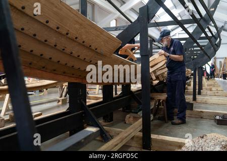 David Steptoe lavora alla replica della nave longship Sutton Hoo, al Longshed di Woodbridge, nel Suffolk. Tre apprendisti falegnami stanno intraprendendo un lavoro di due settimane presso la Sutton Hoo Ship Company, che sta conducendo lavori sulla replica full-size lunga 88 piedi. Data immagine: Venerdì 11 agosto 2023. Foto Stock