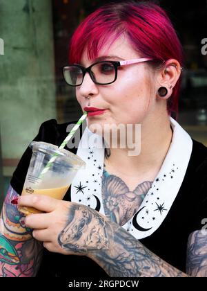 Donna di vent'anni con capelli rossi tinti e tatuaggi con occhiali che beve un caffè ghiacciato fuori da un caffè a Leeds West Yorkshire, Inghilterra Foto Stock