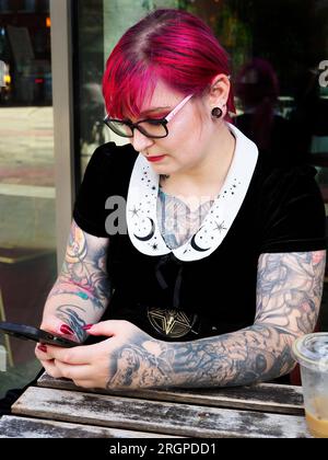 Donna di vent'anni con capelli rossi tinti e tatuaggi che indossa occhiali con uno smartphone a Leeds West Yorkshire in Inghilterra Foto Stock