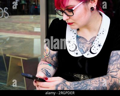 Donna di vent'anni con capelli rossi tinti e tatuaggi che indossa occhiali con uno smartphone a Leeds West Yorkshire in Inghilterra Foto Stock