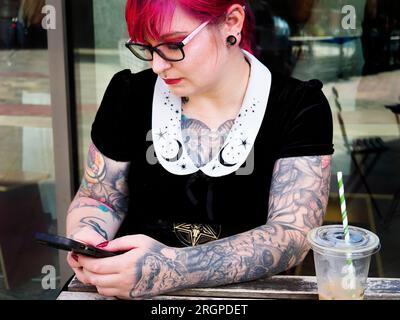 Donna di vent'anni con capelli rossi tinti e tatuaggi che indossa occhiali con uno smartphone a Leeds West Yorkshire in Inghilterra Foto Stock