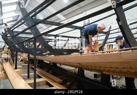 Mike Pratt e David Steptoe lavorano alla replica della nave da diporto Sutton Hoo, al Longshed di Woodbridge, Suffolk. Tre apprendisti falegnami stanno intraprendendo un lavoro di due settimane presso la Sutton Hoo Ship Company, che sta conducendo lavori sulla replica full-size lunga 88 piedi. Data immagine: Venerdì 11 agosto 2023. Foto Stock