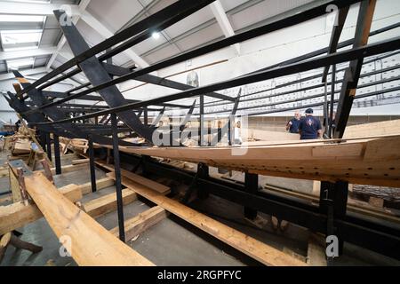 I volontari lavorano alla replica della nave da diporto Sutton Hoo, al Longshed di Woodbridge, Suffolk. Tre apprendisti falegnami stanno intraprendendo un lavoro di due settimane presso la Sutton Hoo Ship Company, che sta conducendo lavori sulla replica full-size lunga 88 piedi. Data immagine: Venerdì 11 agosto 2023. Foto Stock