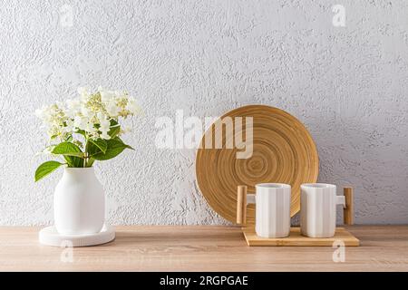 Eleganti tazze in ceramica bianca con manico in legno sul ripiano della cucina e vaso di fiori bianchi. Splendidi interni in stile minimalista Foto Stock