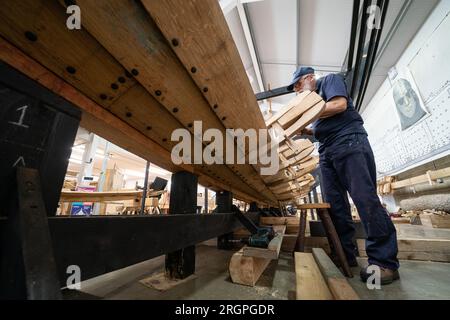 David Steptoe lavora alla replica della nave longship Sutton Hoo, al Longshed di Woodbridge, nel Suffolk. Tre apprendisti falegnami stanno intraprendendo un lavoro di due settimane presso la Sutton Hoo Ship Company, che sta conducendo lavori sulla replica full-size lunga 88 piedi. Data immagine: Venerdì 11 agosto 2023. Foto Stock