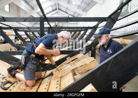 David Steptoe lavora alla replica della nave longship Sutton Hoo, al Longshed di Woodbridge, nel Suffolk. Tre apprendisti falegnami stanno intraprendendo un lavoro di due settimane presso la Sutton Hoo Ship Company, che sta conducendo lavori sulla replica full-size lunga 88 piedi. Data immagine: Venerdì 11 agosto 2023. Foto Stock