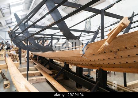 La replica della nave longship Sutton Hoo, al Longshed di Woodbridge, nel Suffolk. Tre apprendisti falegnami stanno intraprendendo un lavoro di due settimane presso la Sutton Hoo Ship Company, che sta conducendo lavori sulla replica full-size lunga 88 piedi. Data immagine: Venerdì 11 agosto 2023. Foto Stock