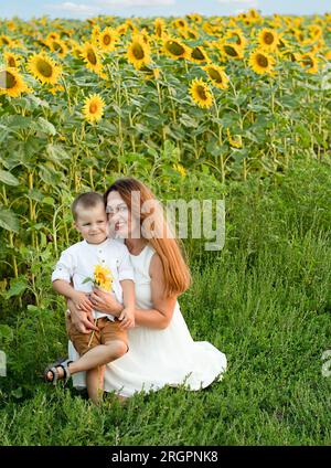 Concetto di famiglia. Una bella donna con un bambino è seduta sull'erba in un campo con girasoli gialli. La madre abbraccia delicatamente suo figlio. Foto Stock
