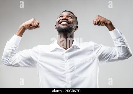 L'uomo nero espelle, le armi alzate, sorridendo per il successo degli sforzi, non per fortuna. Festeggiamo buone notizie, un futuro brillante. Indossa un'elegante camicia bianca come una Foto Stock