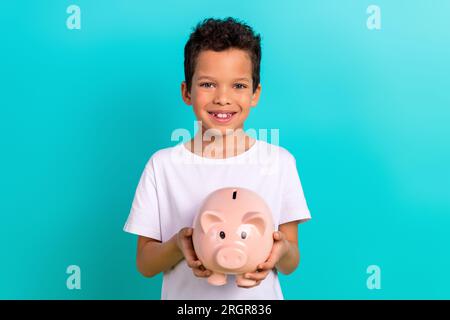 Ritratto di un allegro e carino ragazzo latino che indossa abiti bianchi alla moda tiene l'accumulo di salvadanaio isolato su sfondo di colore ciano Foto Stock