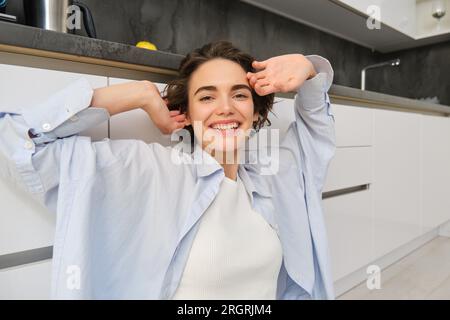 Ritratto ravvicinato di una bella donna sorridente, che tende le mani con il viso contento, si sente stanca, siede sul pavimento della cucina a casa sua Foto Stock