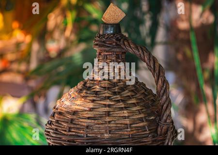 Bottiglia di vino Vintage grande rivestita in vimini. Vecchio Demijohn con sughero ai raggi del tramonto su un tavolo di vetro con tappi di vino sotto il bicchiere. Foto Stock