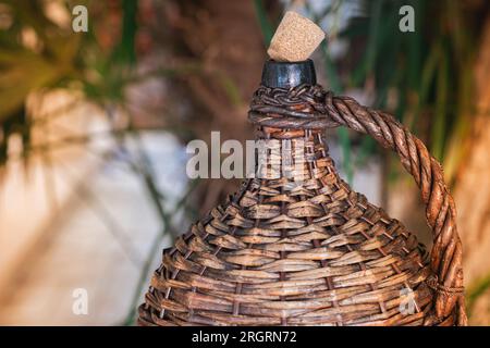 Bottiglia di vino Vintage grande rivestita in vimini. Vecchio Demijohn con sughero ai raggi del tramonto su un tavolo di vetro con tappi di vino sotto il bicchiere. Foto Stock