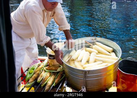 Un venditore prepara e vende con abilità mais cotto e arrosto su una barca a Xochimilco, Messico. L'ampia vasca piena di orecchie fumanti di mais aggiunge un Foto Stock