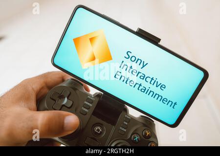 In questa immagine, una persona gioca su un joystick e il logo Sony Interactive Entertainment viene visualizzato sullo schermo di uno smartphone. Foto Stock