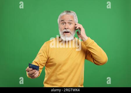 Un uomo anziano scioccato con carta di credito che parla sullo smartphone su sfondo verde. Attenzione - frode Foto Stock