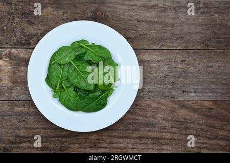 Vista dall'alto e piatta di una pila di foglie di spinaci leggermente bagnate su un piatto bianco su uno sfondo rustico in legno con illuminazione soffusa e spazio per le copie Foto Stock