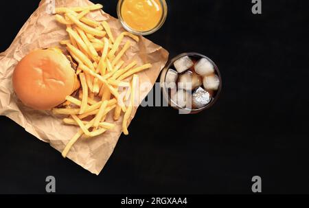 Cheeseburger e patatine fritte su carta marrone e tavolo in legno, vista dall'alto. Hamburger, patatine fritte, cola con ghiaccio in un bicchiere e senape. Fast food Foto Stock