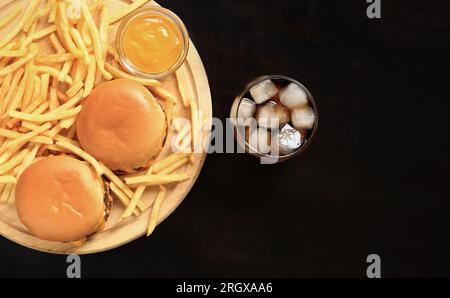 Cheeseburger e patatine fritte su un tagliere, cola con ghiaccio, vista dall'alto, sfondo di legno scuro. Fast food. Hamburger, patatine fritte e senape Foto Stock