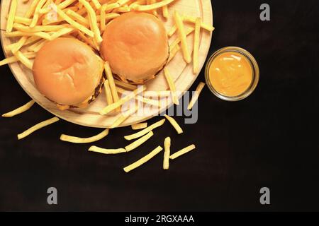 Cheeseburger e patatine fritte su un tagliere, vista dall'alto, sfondo di legno scuro. Fast food. Hamburger, patatine fritte e senape Foto Stock