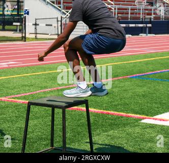 Un ragazzo afro-americano delle scuole superiori che salta e atterra su una scatola di plyo medaglia nera su un campo di erba sintetica verde durante le prove da un lato. Foto Stock