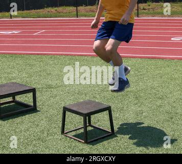 Vista frontale di un ragazzo adolescente in aria che salta su una piccola plyo box nera su un campo di erba sintetica verde durante la pratica. Foto Stock