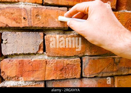 Tiene il gesso bianco della scuola, pronto per scrivere o disegnare su mattoni rossi di un muro danneggiato obsoleto e fatiscente. Foto Stock