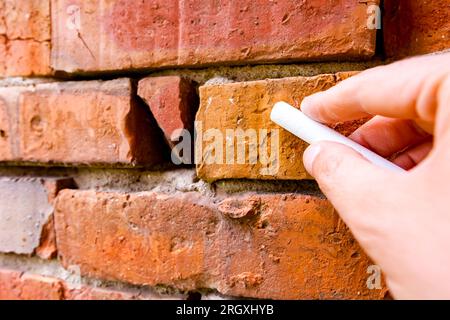 Tiene il gesso bianco della scuola, pronto per scrivere o disegnare su mattoni rossi di un muro danneggiato obsoleto e fatiscente. Foto Stock