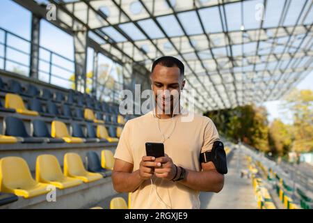 L'atleta maschile utilizza l'app sul telefono dopo la lezione di fitness allo stadio, l'allenatore in cuffie utilizza lo smartphone per ascoltare musica, camminare, sorridere e riposare. Foto Stock