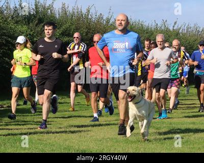 Kesgrave, Suffolk - 12 agosto 2023: 400th Park Run di Kesgrave al Millennium Field. Un uomo che corre con il suo cane tipo labrador. Foto Stock