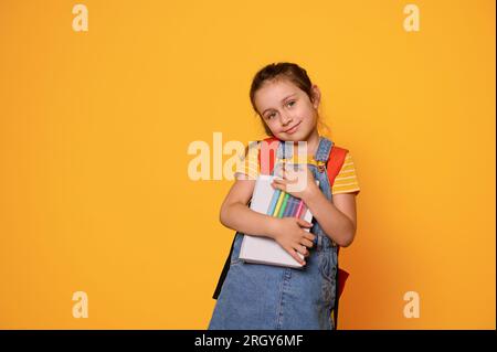 Bellissima bambina con penne in feltro, che porta uno zaino, sorride graziosamente, guarda la fotocamera, isolata su sfondo arancione. Preparazione per t Foto Stock