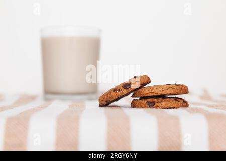 Primo piano di biscotti impilati con gocce di cioccolato e un bicchiere di latte su un tappetino a righe in un tavolo. Attenzione selettiva alla pila di biscotti, ba bianca Foto Stock