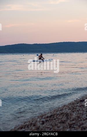 L'uomo dai capelli grigi e dalle barbabietole invecchiate nuota su una linfa in mare al tramonto. Bella e tranquilla pulizia del mare e delle montagne sullo sfondo. Foto Stock