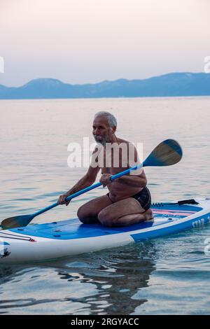 L'uomo dai capelli grigi e dalle barbabietole invecchiate nuota su una linfa in mare al tramonto. Bella e tranquilla pulizia del mare e delle montagne sullo sfondo. Foto Stock