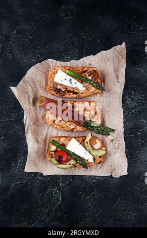 Pane tostato fatto in casa servito con asparagi verdi avvolti in pancetta, formaggio, fette di avocado, ciliegia di pomodoro alla griglia e noci su fondo di cemento scuro Foto Stock