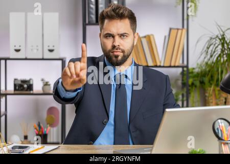 Un giovane uomo d'affari caucasico che lavora su un computer portatile scuote il dito e dice di no fai attenzione a rimproverare e dare consigli per evitare errori di pericolo segno di disapprovazione in ufficio. Uomo libero professionista sicuro di sé Foto Stock