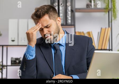 Uomo d'affari caucasico stanco malato che soffre di mal di testa, problemi di tensione e emicrania, stress da casa ufficio scrivania sul posto di lavoro. Il lavoratore mediatore freelance barbuto lavora su notebook. Lavoro eccessivo Foto Stock