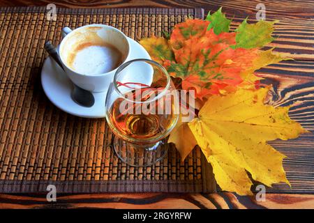 la foto mostra una natura morta autunnale con una tazza di caffè, un bicchiere di brandy e foglie luminose. Foto Stock