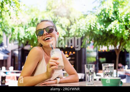 Una giovane bruna felice seduta nel caffè all'aperto che beve caffè ghiacciato e ride. Foto Stock
