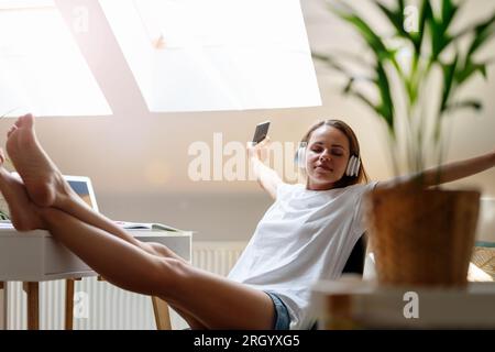 la giovane donna siede con i piedi in alto sul tavolo ascolta la musica utilizzando le cuffie wireless e lo smartphone, ama la musica, allunga le braccia e bo Foto Stock