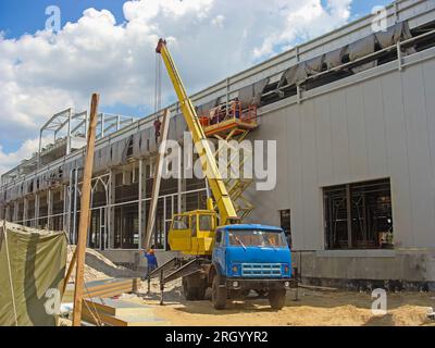 Costruzione di un grande magazzino logistico. Un cantiere con una gru che solleva un pannello sandwich per l'installazione Foto Stock