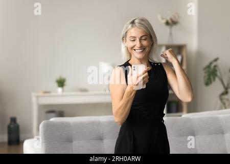 Bella donna bionda matura allegra che legge un messaggio entusiasmante sullo smartphone Foto Stock